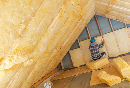 A worker is installing insulation in the sloped roof of a residential renovation project. The bright yellow insulation panels are being fitted to improve energy efficiency. The setting is partially completed with debris around.の写真素材