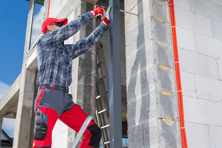 A construction worker in a plaid shirt and red pants works diligently on a building site. He is using tools to install house gutter.の写真素材