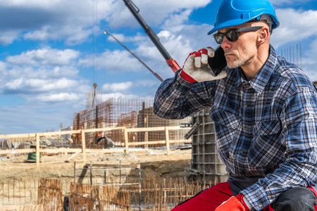 A construction worker in a blue helmet and protective gear is sitting on a site while talking on his smartphone. Behind him, cranes and construction materials indicate ongoing building activities under a cloudy sky.の写真素材