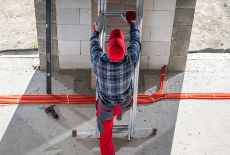 A construction worker in a red hat and gloves climbs a metal ladder to reach a brick wall. The worker wears a plaid shirt and red pants, focusing on the task at hand.の写真素材