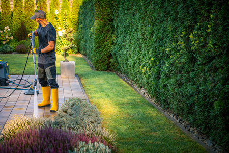A man dressed in work attire and yellow boots trims the grass in a well-maintained garden. The lush greenery and vibrant flowers create a tranquil atmosphere in the afternoon sun, showing his dedication to outdoor care.の写真素材