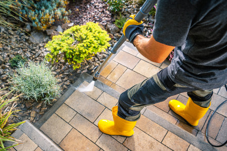A person is using a pressure washer to clean stone steps in an outdoor setting. They are wearing bright yellow boots and black gloves while spraying water on the stairs.の写真素材