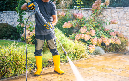 A person wearing yellow boots and gloves operates a pressure washer while cleaning a paved patio in a lush garden. Colorful flowers and greenery surround the area, showing a serene outdoor environment.の写真素材