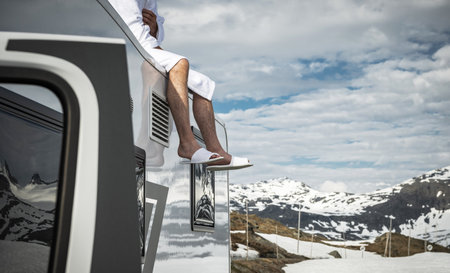 A person wearing a bathrobe and slippers sits atop a camper in a remote mountainous area. Snow-covered peaks surround the landscape under a cloudy sky, creating a serene and peaceful atmosphere.の写真素材