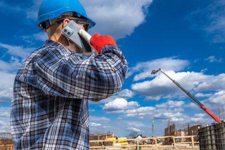 A construction worker wearing a blue hard hat and red gloves stands on a building site, talking on his phone.の写真素材