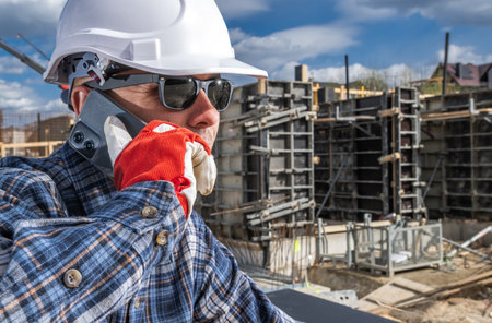 A construction worker in a hard hat and gloves speaks on the phone. He wears sunglasses and a plaid shirt, with construction frames set against a blue sky and scattered clouds. His focused expression suggests coordinating a task.の写真素材