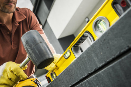 A construction worker is using a yellow level tool to ensure materials are properly aligned. Wearing gloves and focused on the task, he taps gently with a mallet to adjust the position of the materials outside a building.の写真素材