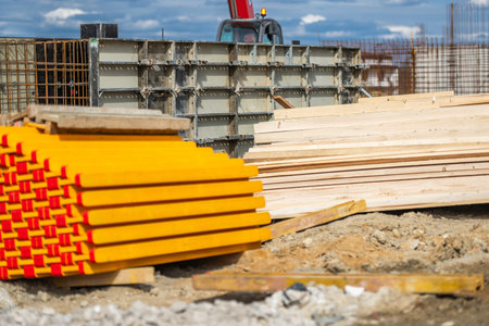 Brightly colored lumber is stacked neatly on the ground at a construction site. Adjacent concrete forms are set up for building activity under a partly cloudy sky. Heavy machinery is visible in the background.の写真素材