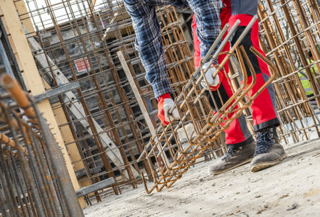A construction worker in a plaid shirt and red pants is bending down to lift a large piece of steel...