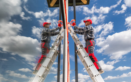 A worker ascends a ladder to perform maintenance on a structure under a bright sky. The reflection of the worker can be seen on the surface nearby, highlighting the activity against the dynamic cloud backdrop.の写真素材