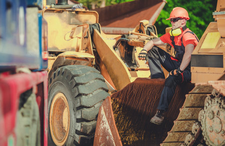 A construction worker takes a break sitting on heavy machinery at a construction site. Wearing a hard hat and safety gear, he enjoys the surroundings while being surrounded by large equipment on a sunny day.の写真素材