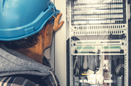 An experienced technician wearing a blue hard hat inspects electronic equipment in a server room. The scene captures a busy work environment with servers and cables visible, illustrating high-tech operations.の写真素材