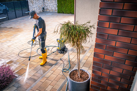 A man stands on a stone patio wearing yellow boots and a cap, using a pressure washer to clean the surface. The area is well-maintained, featuring decorative plants and a vibrant green backdrop.の写真素材