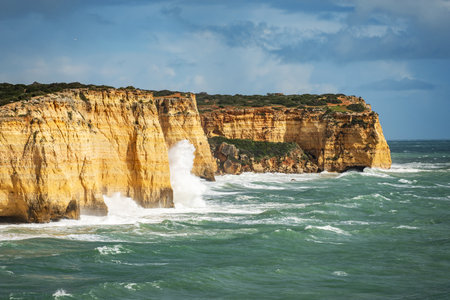 Along the coastline, waves violently crash against dramatic yellow cliffs, sending up sprays of white foam. The ocean is a deep blue-green, contrasting sharply with the warm tones of the rocky outcrops.の写真素材