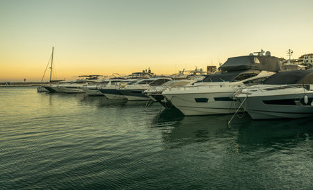 A stunning view captures the calm waters of a marina filled with luxury yachts, peacefully moored as the sun sets. The golden hues of the sky reflect on the water, creating a serene atmosphere.の写真素材