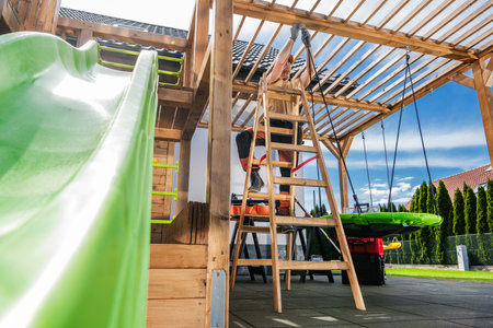 A child enjoys sliding down a bright green slide while a parent works on constructing a wooden play...
