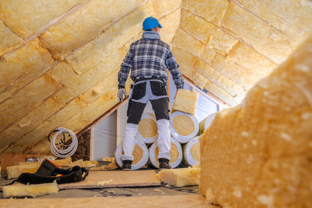 A worker wearing safety gear stands in an attic surrounded by rolls of insulation material. Sunlight filters through the roof, illuminating the unfinished space. The worker carefully inspects the area, preparing to enhance the home's energy efficiency by installing insulation in the ceiling.の写真素材