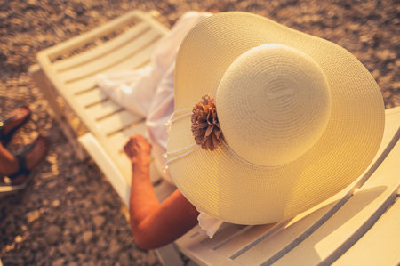 A woman relaxes on a sun lounger at the beach, wearing a wide-brimmed hat adorned with a floral detail. The warm sunlight casts a glow on the sandy surroundings.の写真素材
