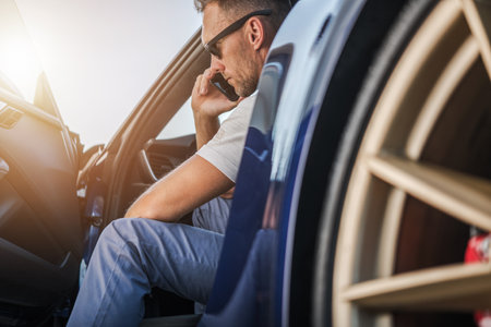 A man dressed casually sits in the driver's seat of a car, engaged in a phone conversation as the...
