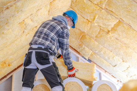 A construction worker in a blue helmet and checkered shirt is installing insulation in a home's attic during daylight. Safety gear is worn for protection.の写真素材