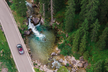 A convertible car travels along a winding road next to a vibrant stream, surrounded by tall trees...