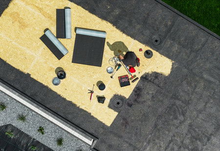 A worker repairs a roof in a sunny environment using various tools and materials. The roof shows sections being covered while others remain bare.の写真素材