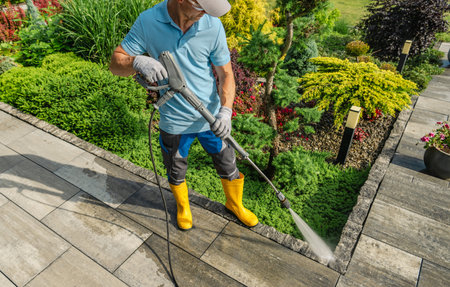 A man in yellow boots cleans a stone pathway using a pressure washer, surrounded by lush green plants and blooming flowers on a sunny day.の写真素材