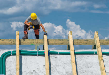 A worker carefully places wooden beams on a roof structure while standing on scaffolding. Bright...