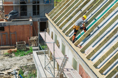 A construction worker carefully places roof beams on a residential building while scaffolding is...