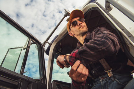 A truck driver adjusts his watch as he leans out of his vehicle's cabin. He wears sunglasses and a cap, surrounded by a rural landscape and cloudy skies.の写真素材