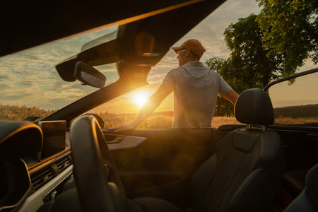 A man stands beside a convertible car parked in a field, gazing at the stunning sunset on a clear evening. The warm light casts a golden glow.の写真素材