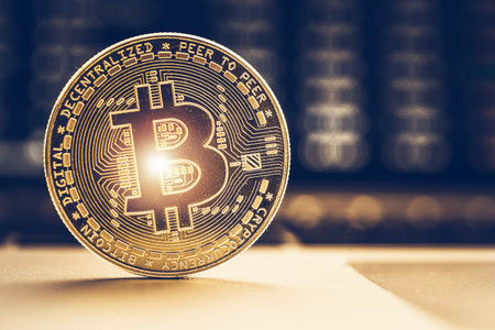 A Bitcoin coin stands upright on a table, showing its intricate design and gold color, while a...