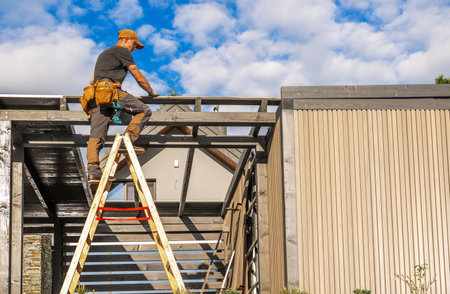 A construction worker balances on a ladder, carefully installing wooden beams on a building extension. The job takes place on a sunny afternoon with few clouds.の写真素材
