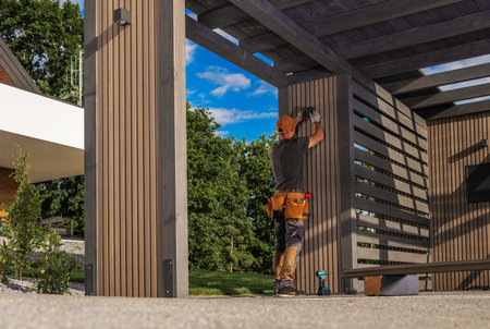 A construction worker is installing a light fixture on wooden panels to a frame outside, showcasing...