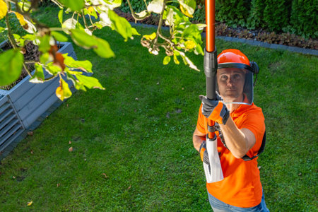 A man in an orange shirt uses a long pole saw to trim tree branches in a green backyard, surrounded...