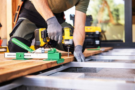 A skilled craftsman uses a drill to secure wooden planks on a deck frame while wearing gloves. Tools...