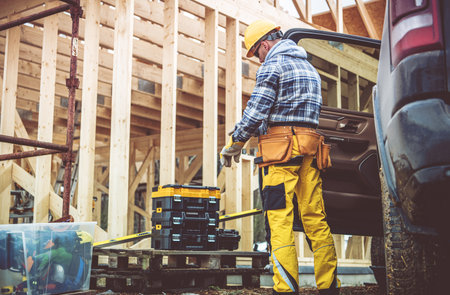 A construction worker in bright yellow attire is measuring tools at a building site. The wooden...