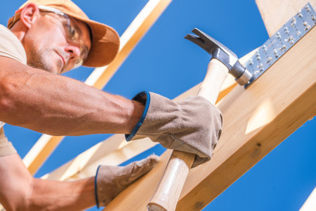 A carpenter hammers a wooden beam while constructing a framework. The worker is focused, wearing gloves, with a bright blue sky in the background.の写真素材