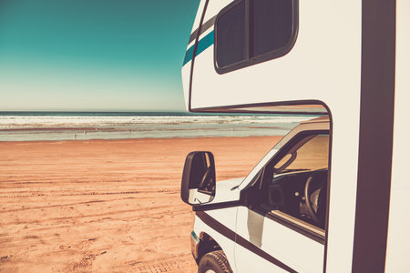 A camper van parked on a sandy beach with ocean waves in the background under a clear blue sky. Perfect weather for a seaside getaway.の写真素材