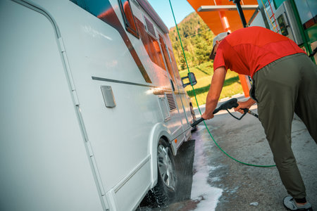 A man carefully washes his camper van using a pressure washer at a service station, surrounded by green hills and clear skies, on a sunny afternoon.の写真素材