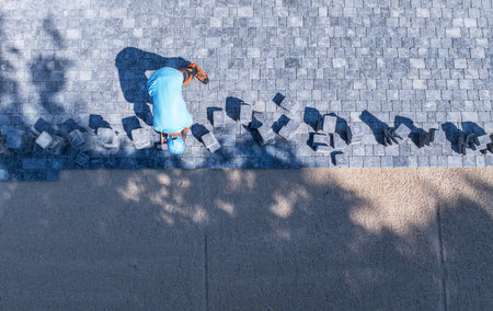 A worker carefully places stone pavers on a pathway, ensuring precision and alignment under the warm sun. Shadows from trees add depth to the scene.の写真素材