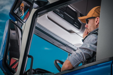 A truck driver wearing sunglasses and a cap is talking on his phone inside the cabin, looking...