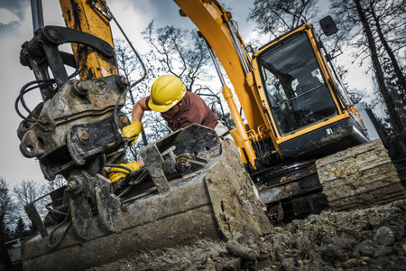 An excavator operator in a hard hat and gloves adjusts machinery in muddy conditions at a construction site surrounded by trees during daylight hours.の写真素材