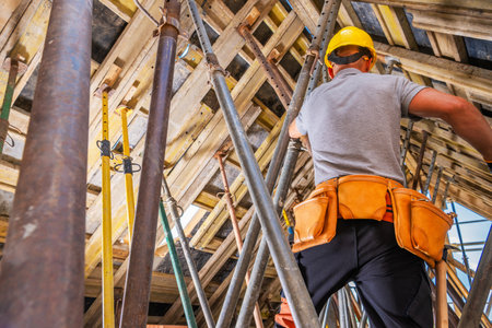 A construction worker wearing a helmet and harness assembles scaffolding at a high-rise site,...