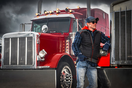 A truck driver leans against a red semi truck under a cloudy sky. The scene shows the driver in casual attire, enjoying a break during the day.の写真素材