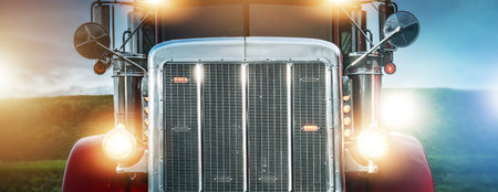 A large truck stands in a field with headlights shining brightly as dusk approaches. The sky shows...