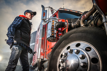 Man adjusts a wrench on a large red truck during daytime at a worksite. Smoke rises in the background.の写真素材