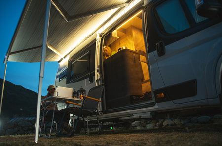 People are relaxing outside a camper with light coming from inside at night. One person is sitting...