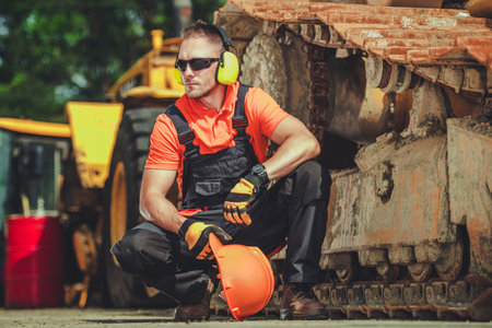 A worker in an orange shirt and black overalls squats next to a large machine on a construction...