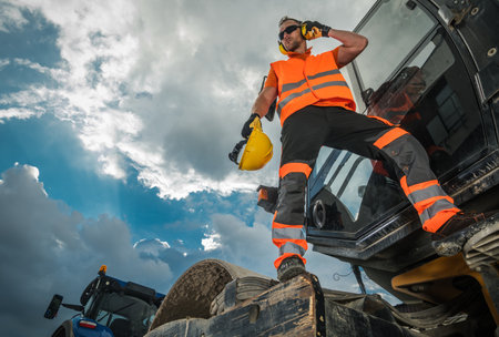 A worker wearing safety gear talks on the phone while standing on heavy machinery at a construction...